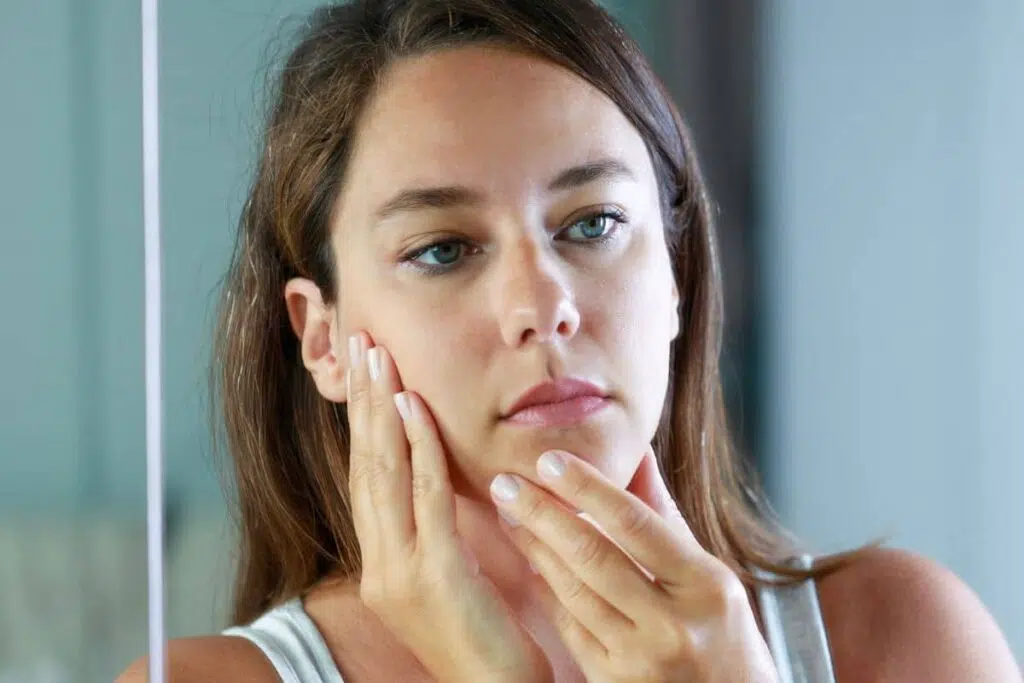 Concerned woman touching her face in mirror, assessing volume loss and skin laxity after weight loss