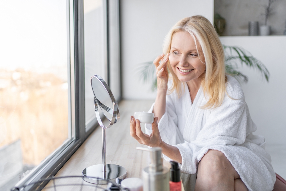 Woman applying face cream near window and mirror, representing PRF EZ Gel recovery and gentle post-treatment skincare routine