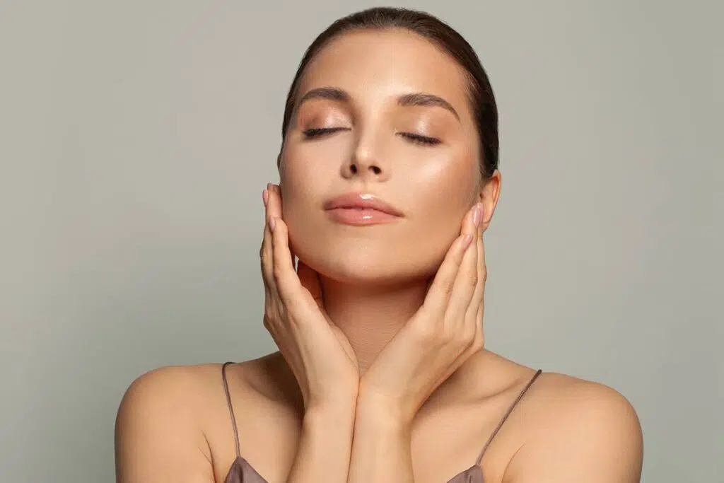 A close-up view of a smiling woman with clear, luminous skin looking up and to the side while holding her hand to her jawline.