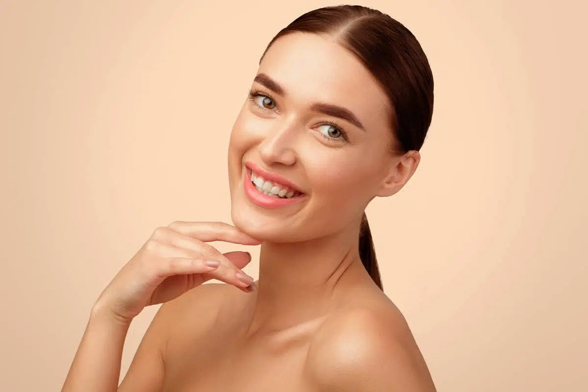 A front portrait of a smiling woman with clear skin tilting her head slightly and resting a finger gently under her well-defined jawline.