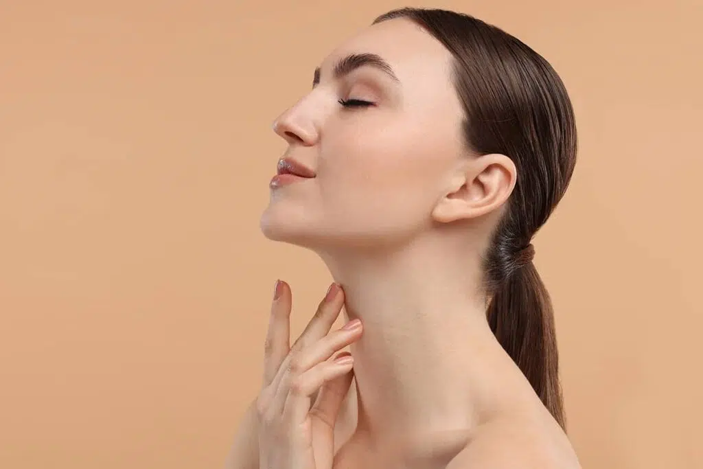 A side profile view of a brunette woman with a ponytail tilting her chin up with eyes closed while touching her neck against a beige background.